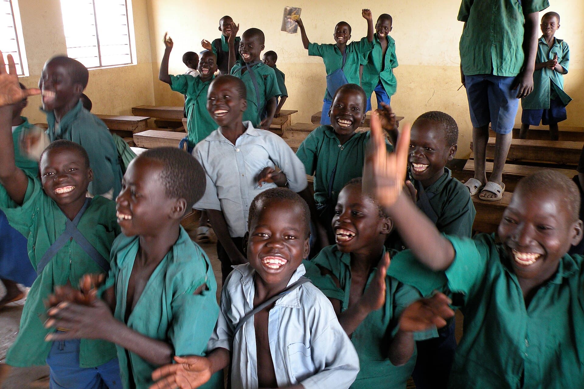 Pupils in green school uniforms smiling and raising their hands in a Ugandan classroom